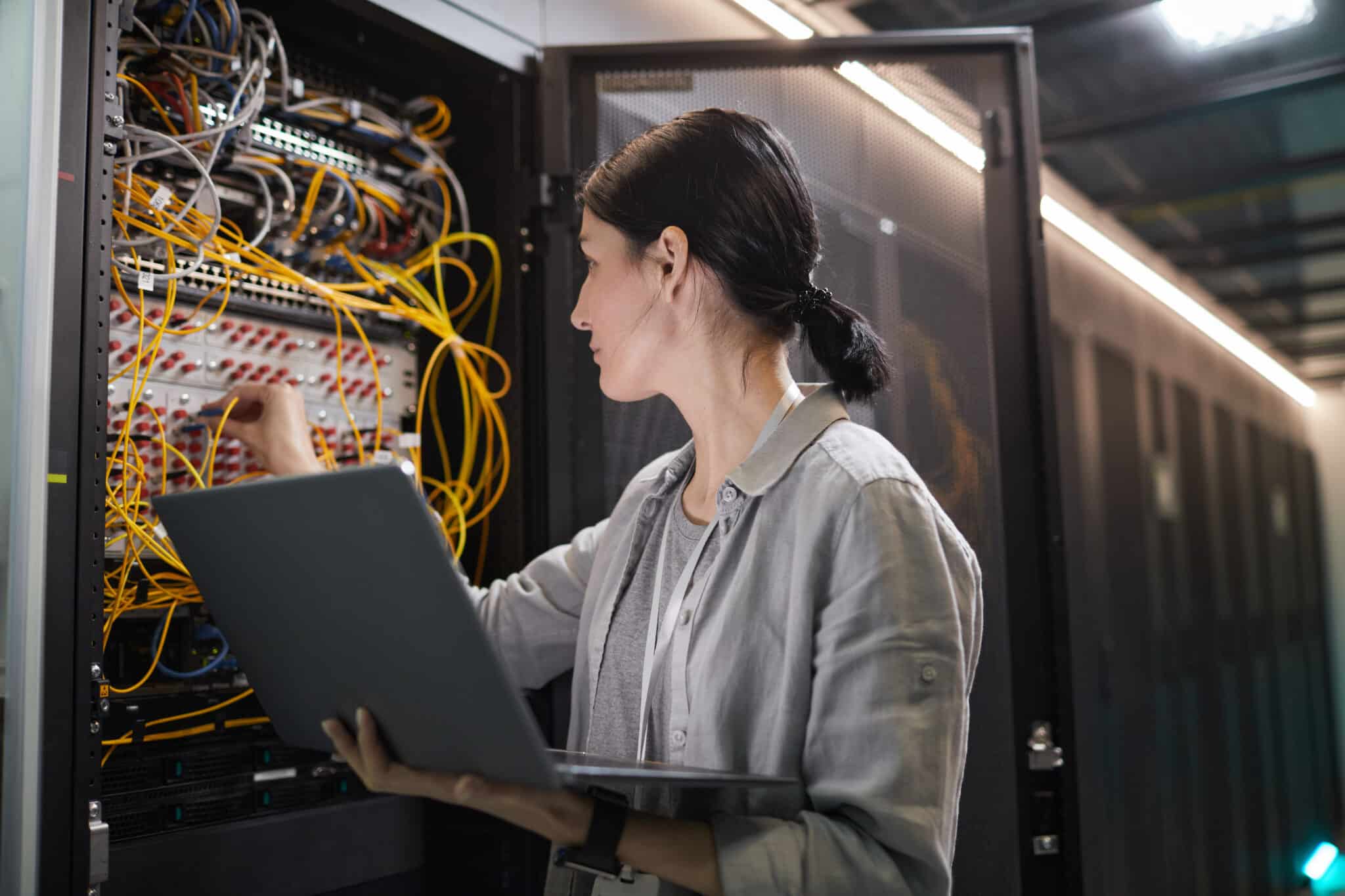 Woman working in front of a networking cabinet while holding a laptop.